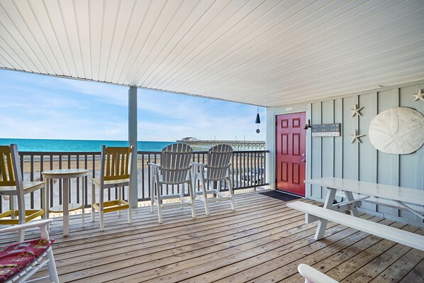 Una veranda coperta con pavimento in legno, poltrone Adirondack bianche e gialle, una porta rossa e vista sull'oceano.