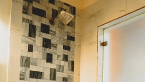 A bathroom with a checkered tile wall, a towel rack with a white towel, a showerhead, and a frosted glass door. - JK Suites (Gurugram)