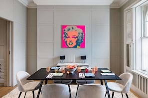 Uma área de refeições com uma mesa posta para quatro pessoas, dois alto-falantes fixados na parede e um grande quadro de arte pop.
