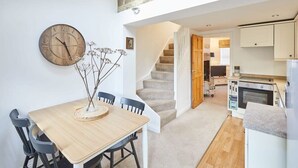 A dining area with a wooden table and chairs, a wall clock, and a vase with dried flowers. A staircase leads to a kitchen area with modern appliances. - Host & Stay | Waterlily Cottage (Stokesley)