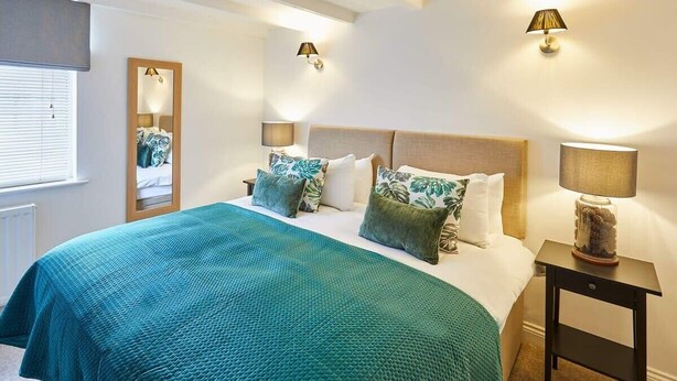 A neatly arranged bedroom with a large bed, two bedside lamps, a mirror, and a window with blinds. - Host & Stay | Waterlily Cottage (Stokesley)