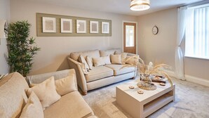 Un salon avec un canapé beige, une table basse et une grande plante. Des tableaux encadrés ornent le mur, et une fenêtre est agrémentée de rideaux.