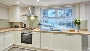 Une cuisine moderne avec des meubles blancs, une hotte en acier inoxydable, un four encastré, un évier à deux bacs et une fenêtre avec des stores.