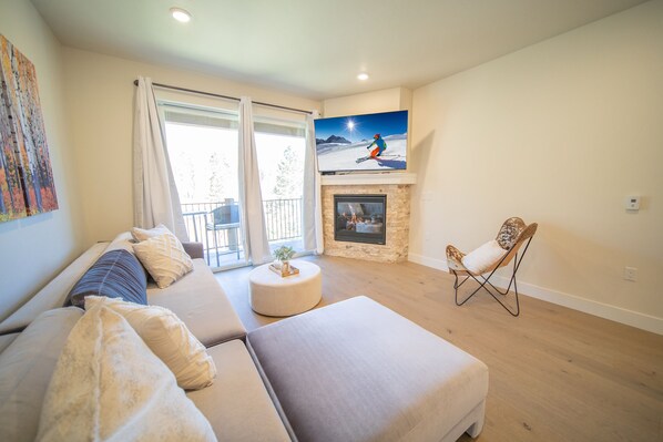 A living room with a sofa, ottoman, and a chair. There's a fireplace with a painting above it. - Modern Keystone Condo | Lake Fun + Hot Tubs (Keystone)