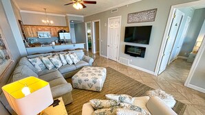 A spacious living area with a grey sectional sofa, a white ottoman, and a yellow lamp. A flat-screen TV is mounted on the wall, and there is a fireplace below it. - Elegant, Stunning, Beach Front Condo; New to Rental Market! Oversized Balcony (Panama City Beach)