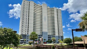 A tall, multi-story hotel with a central vertical feature, surrounded by greenery and a pedestrian bridge. - Elegant, Stunning, Beach Front Condo; New to Rental Market! Oversized Balcony (Panama City Beach)