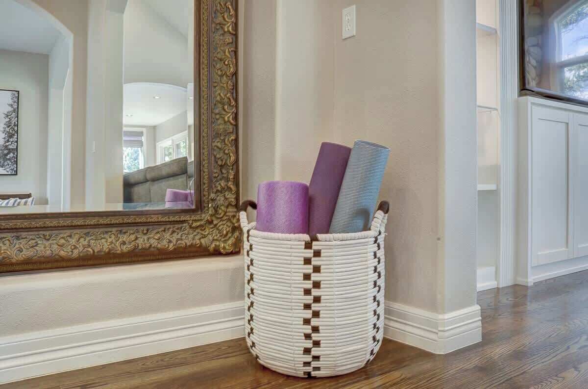 A corner of a room with a white wicker basket containing rolled-up mats, a large ornate mirror, and a wooden floor.