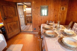 Een houten interieur met een eettafel voor vier personen, een keuken zichtbaar door een open deur, en een klein raam.
