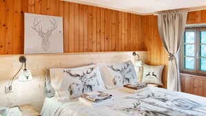 Een kamer met houten lambrisering, een bed met beddengoed met hertenmotieven, een wandlamp en een raam met gordijnen.