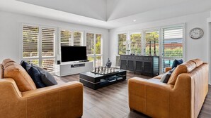 A spacious living room with a large flat-screen TV, leather sofas, a coffee table, and a view of a balcony with greenery. - Hamptons Resort: 3  BR, 2  BA House in Sorrento, Sleeps 6 (Sorrento)