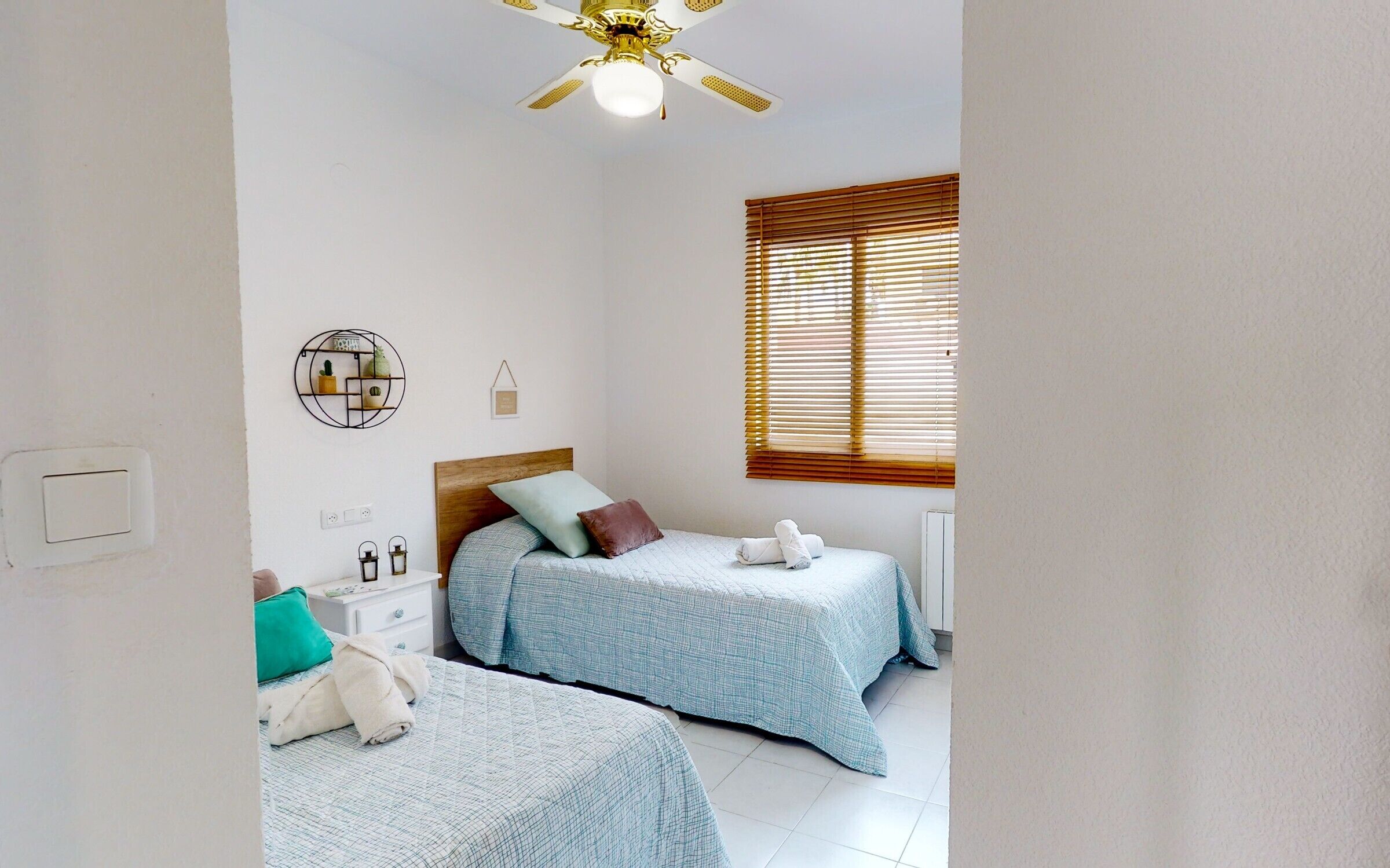A bedroom with two beds, a ceiling fan, a window with blinds, a bedside table with a lamp, and a wall-mounted shelf.