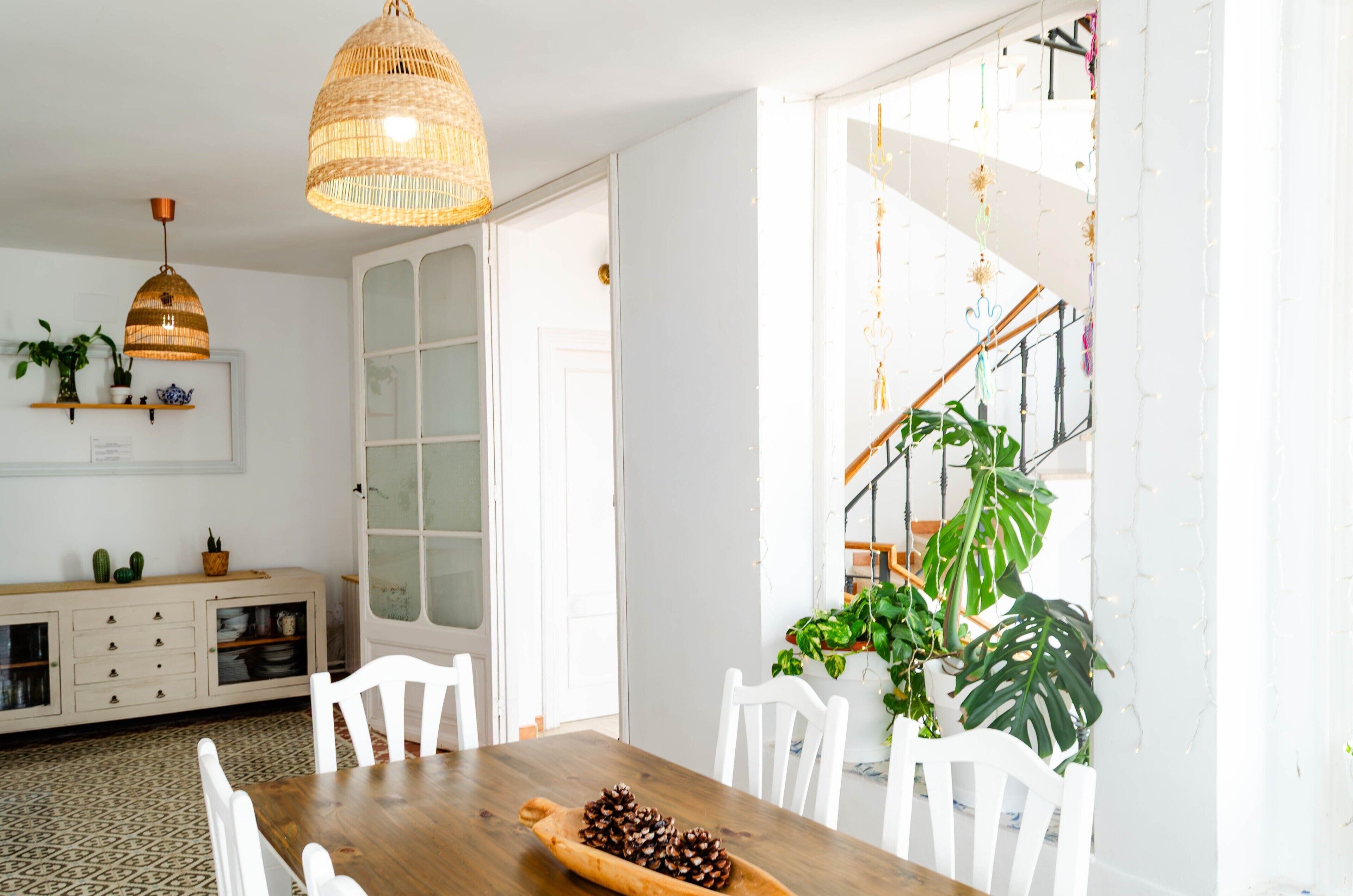 A dining area with a wooden table, white chairs, a woven pendant light, and a staircase with string lights.