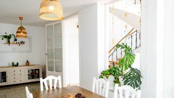 A dining area with a wooden table, white chairs, a woven pendant light, and a staircase with string lights.