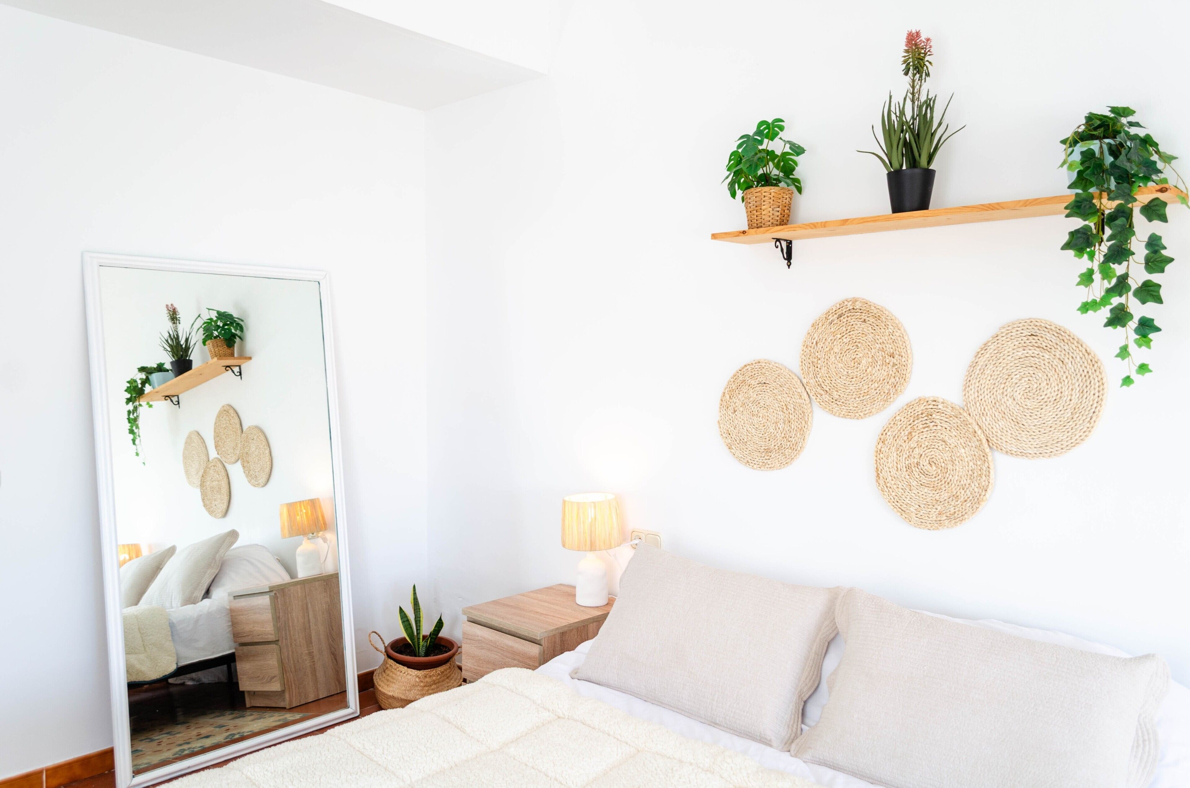 A bedroom with a bed, bedside table, lamp, mirror, and wall-mounted shelves with plants and woven placemats.
