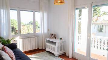 A room with a sofa, a small white console table, a wicker pendant light, and a view of greenery outside.
