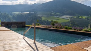 Una piscina con muelle de madera y baranda metálica, rodeada de un jardín ajardinado con flores y vistas a colinas onduladas.