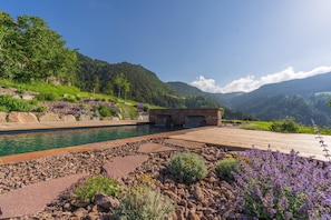 A pool with a wooden deck, surrounded by rocks and plants, with mountains in the background. - Apartment "Spornberg Mountain Living Nordberg" with Mountain View (Renon)