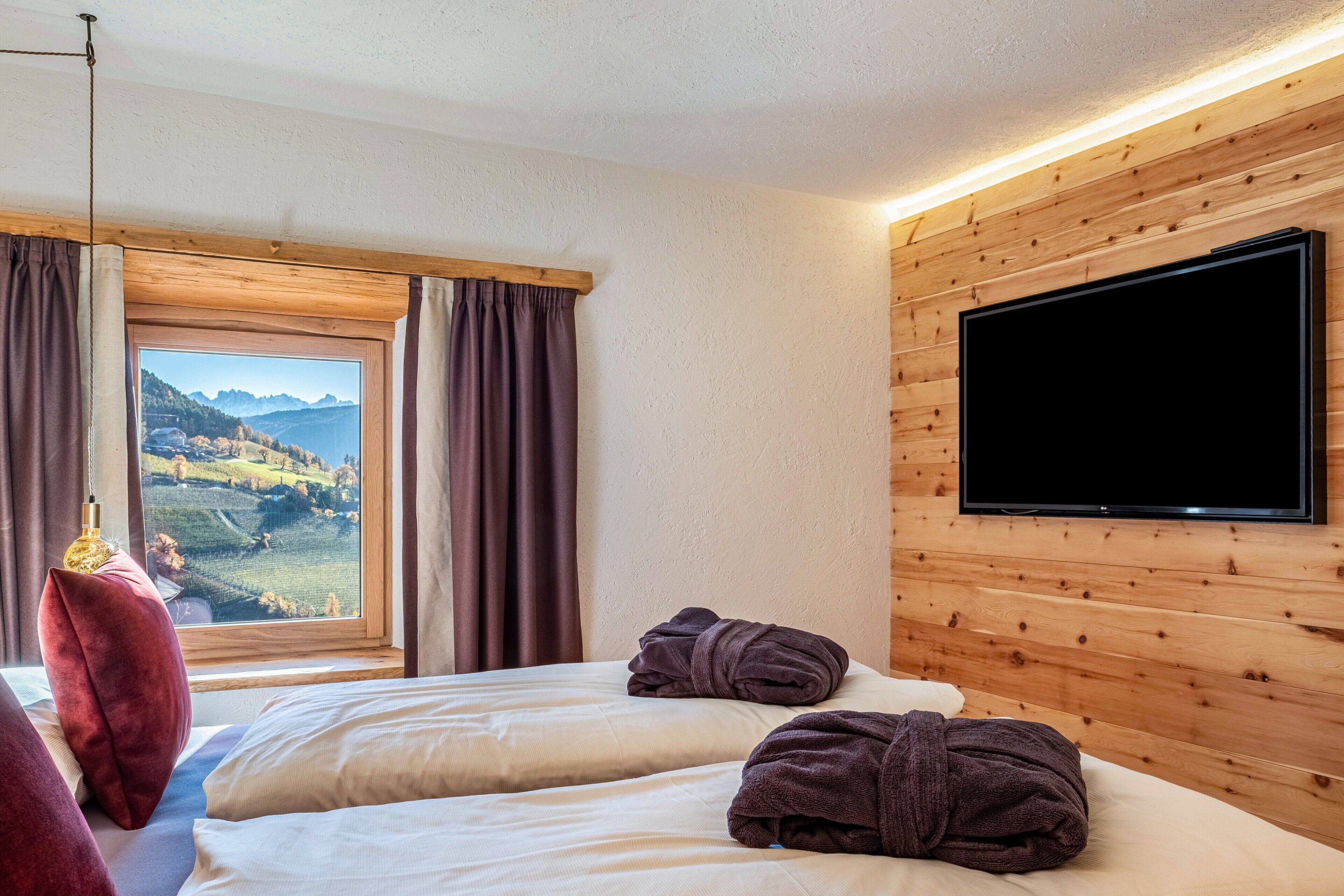 A hotel room with a bed, a TV, a window with curtains, and a view of a mountainous landscape.