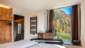 A bedroom with two beds, a wooden wardrobe, a black towel rack, and a view of a forested hillside. - Apartment "Spornberg Mountain Living Ostberg" with Mountain View (Renon)
