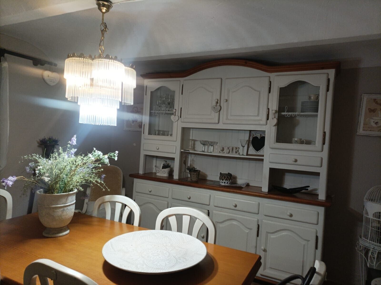 Een eetruimte met een houten tafel, witte stoelen, een kroonluchter en een witte kast met glazen deuren.