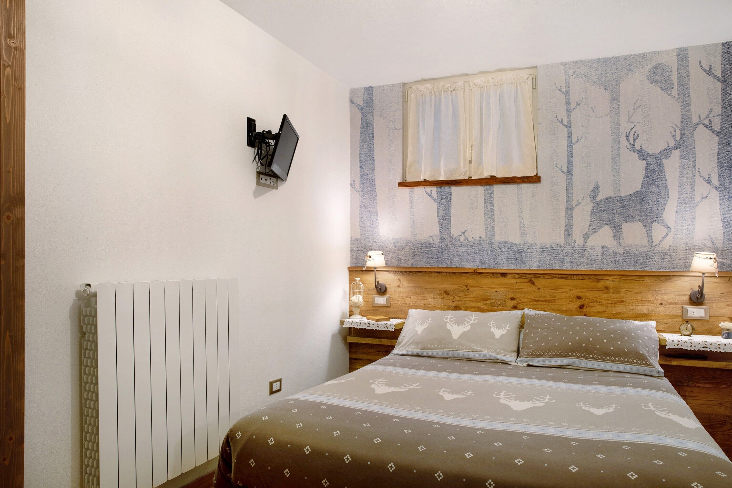 Ein Schlafzimmer mit Bett, Wandfernseher, Fenster mit Vorhängen und Heizkörper.