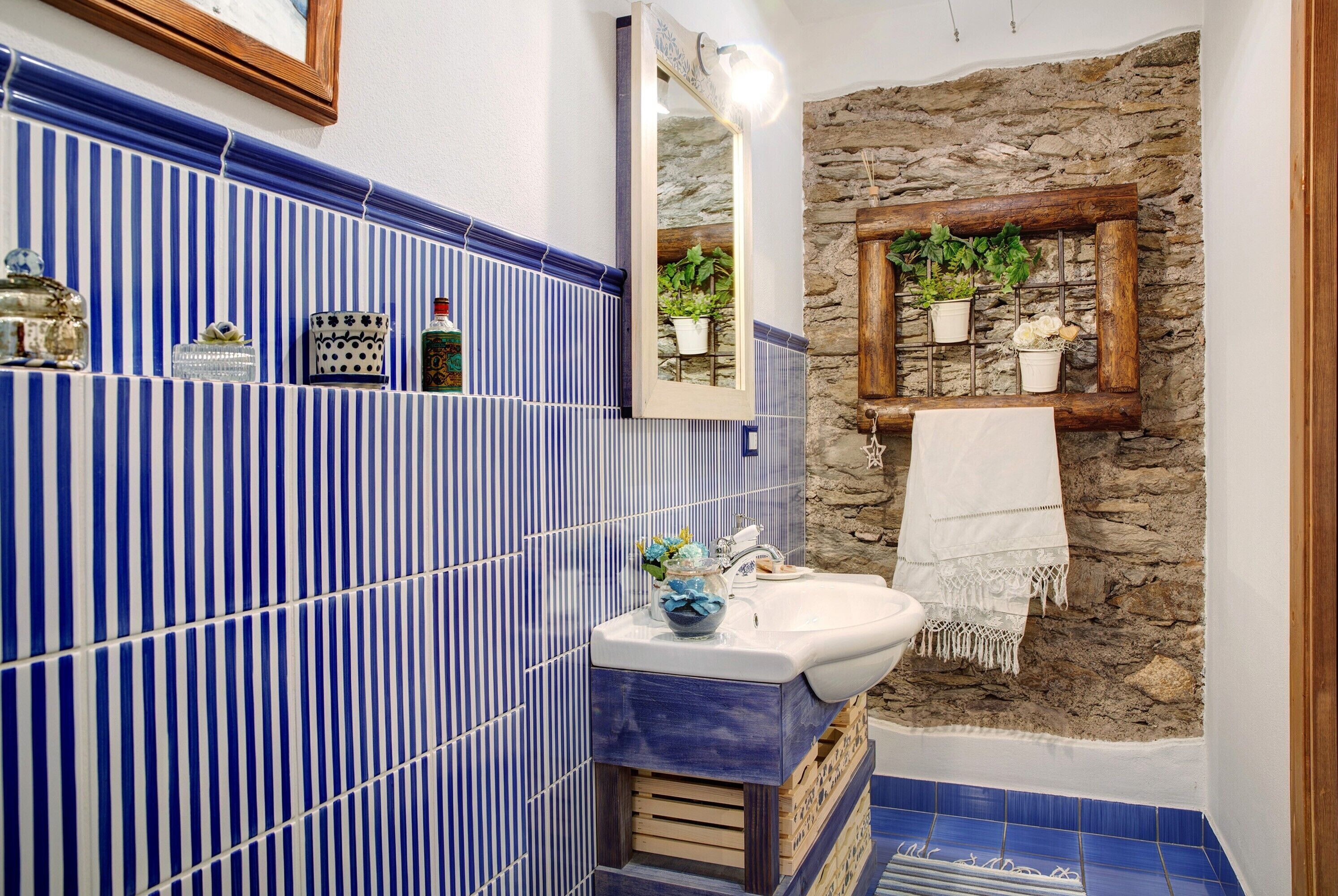 A bathroom with a blue-striped tile wall, a stone accent wall, a wooden shelf with plants, a white sink, and a mirror.
