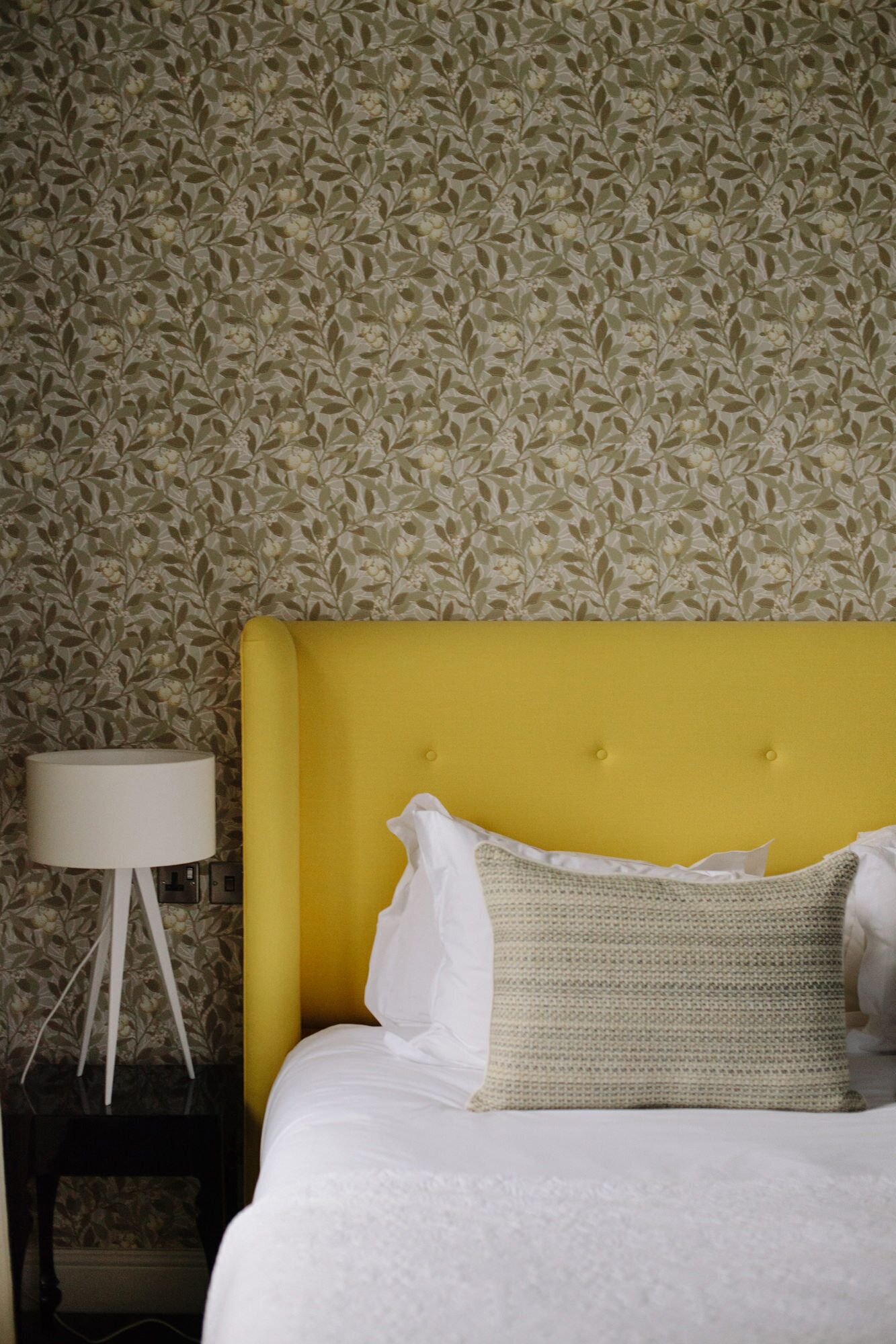 A bed with a yellow headboard, white bedding, and a textured pillow. A white lamp is on a side table against a floral wallpaper background.