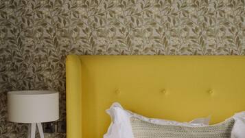 A bed with a yellow headboard, white bedding, and a textured pillow. A white lamp is on a side table against a floral wallpaper background.