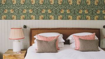 Una cama bien hecha con dos almohadones estampados, una lámpara de noche y una pared con estampado floral.