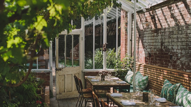 Un patio al aire libre con mesas y bancos, rodeado de vegetación y muros de ladrillo.