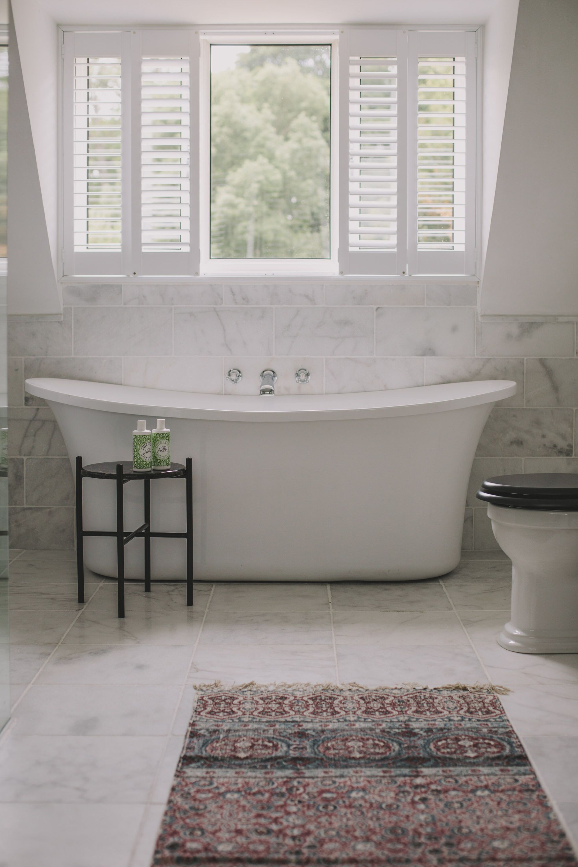 A bathroom with a freestanding bathtub, a window with shutters, and a patterned rug on the floor.