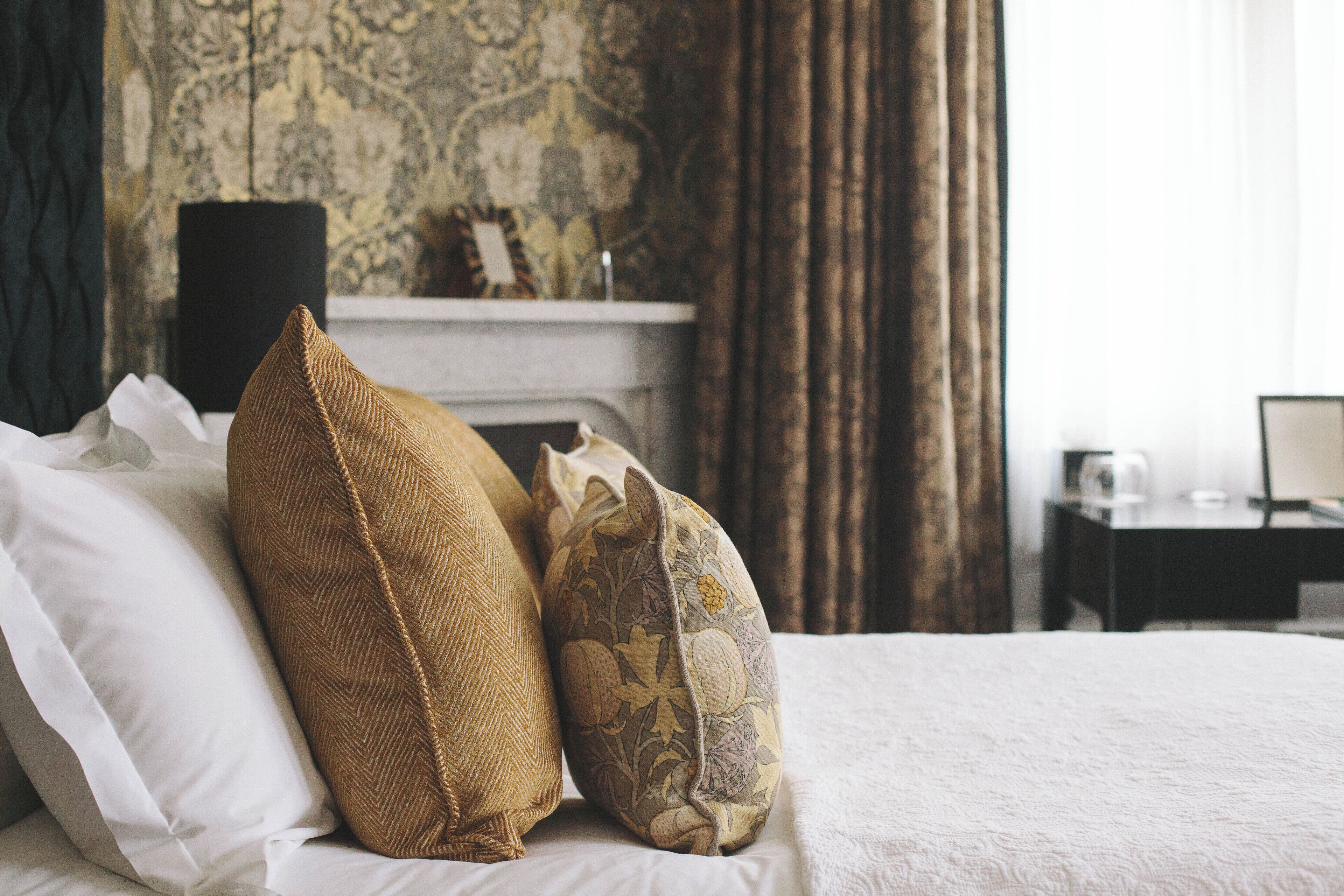 A neatly made bed with white and patterned pillows, a fireplace, and a desk with a lamp in a room with patterned wallpaper.