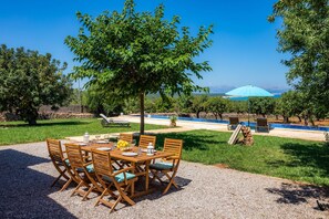 A dining area with a table set for six, surrounded by chairs, a tree, and a view of a body of water. - Finca "Can Jaumi" with Sea Views, Pool and Wi-Fi (Artà)