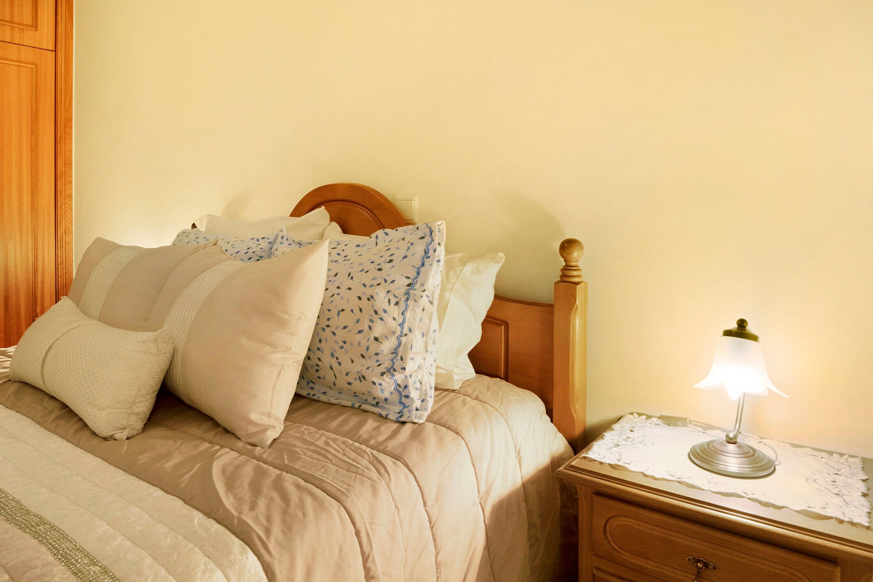 Un letto rifatto con cura, con una testiera in legno, un comodino con una lampada e un piccolo tavolo di legno.