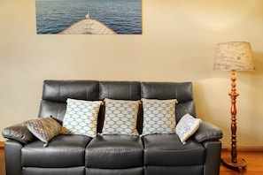 A black leather sofa with patterned cushions, a wooden floor lamp, and a maritime-themed painting on the wall.