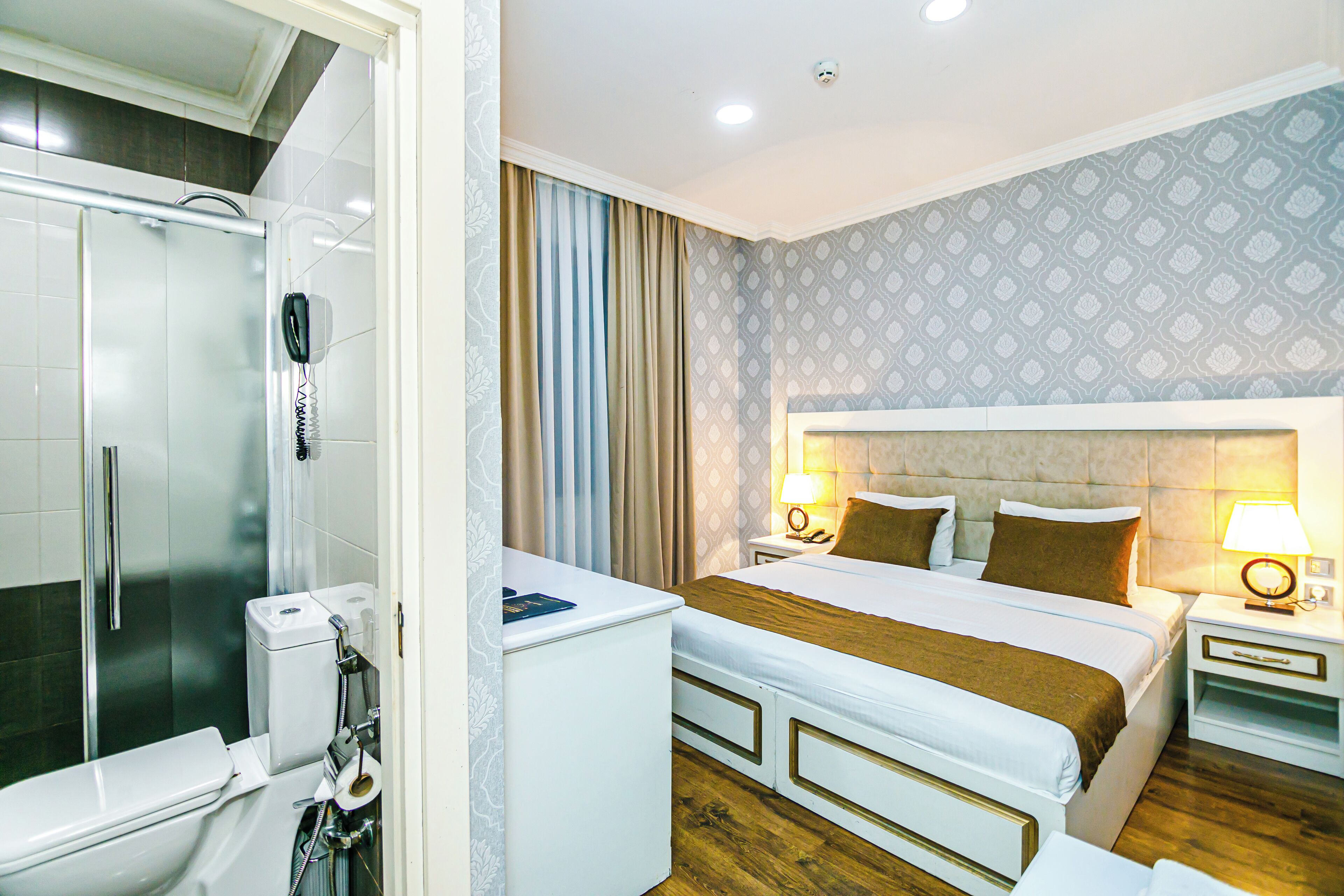 A hotel room with a shower, toilet, bed, bedside tables, and a patterned wall.