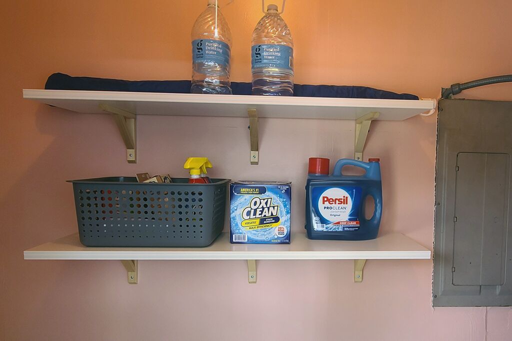A shelf with two water bottles, a laundry basket, a box of OxiClean, and a bottle of Persil ProClean.