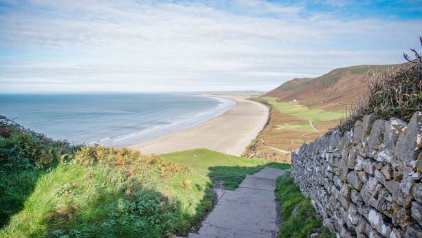 A coastal pathway leading to a beach with a stone wall on one side and a grassy hill on the other. - Old School Cottage Ship Farm Rhossili (Swansea)