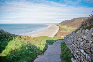 Un sentier côtier mène à une plage, avec un mur de pierres d’un côté et une colline herbeuse de l’autre.