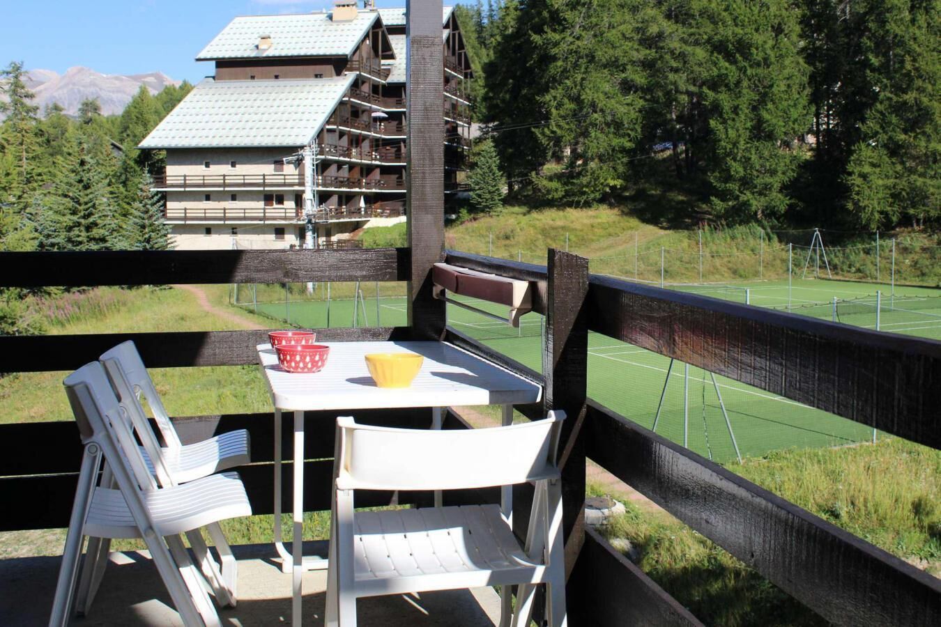 Une terrasse en bois avec une table et des chaises donnant sur un court de tennis, avec un bâtiment en arrière-plan.