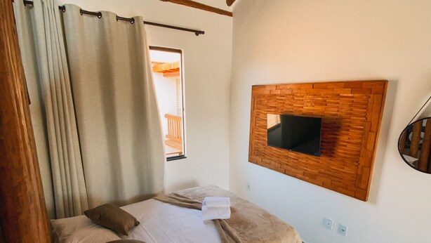 A bedroom with a bed, curtains, a wooden ceiling, a mounted TV, and a window.