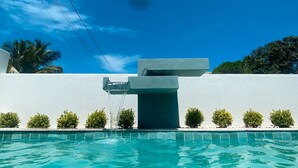 A swimming pool with a modern water feature, surrounded by a white wall and small bushes.