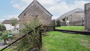 A stone building with a triangular roof, surrounded by a well-maintained garden with flowering plants and a wooden fence.