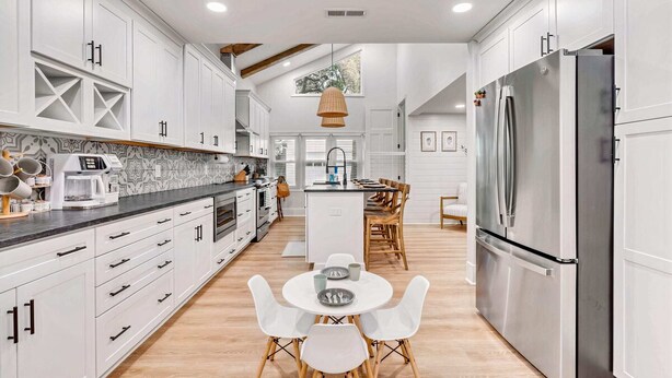 A modern kitchen with white cabinetry, a stainless steel refrigerator, and a dining area with white chairs. - Luxury Isle of Palms Home Steps to Beach with Pool (Isle of Palms)