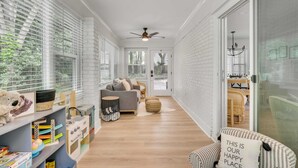 A bright, well-lit hallway with a grey sofa, a small round ottoman, and a bookshelf with toys and books. - Luxury Isle of Palms Home Steps to Beach with Pool (Isle of Palms)
