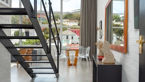 Un interior moderno con una mesa de vidrio, sillas transparentes, una escalera de metal negro y vistas a edificios a través de grandes ventanales.