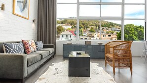 Una sala de estar moderna con un sofá gris, una mesa de centro negra, un sillón de mimbre y un ventanal con vistas a un pueblo en la ladera.