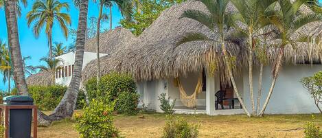 A thatched-roof house with a porch, surrounded by palm trees and a garden with stepping stones.