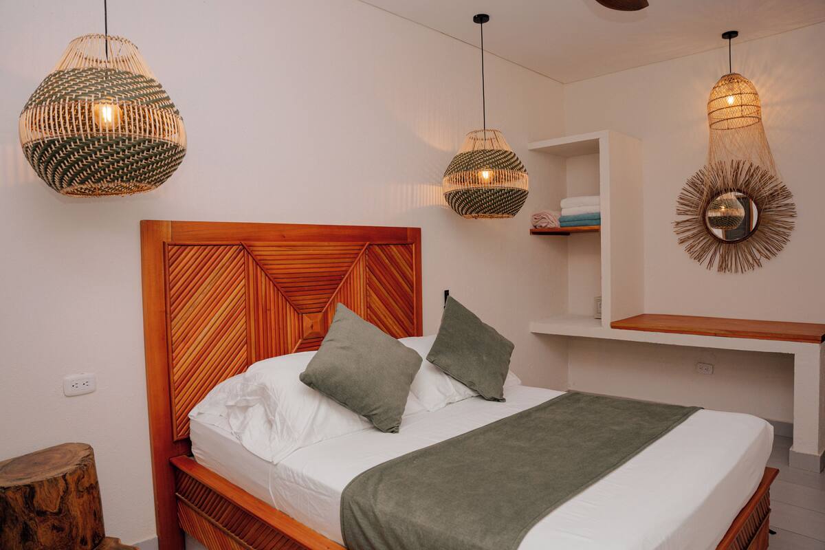A neatly made bed with a wooden headboard, two pillows, and a green throw blanket. There are three woven pendant lights hanging above the bed.