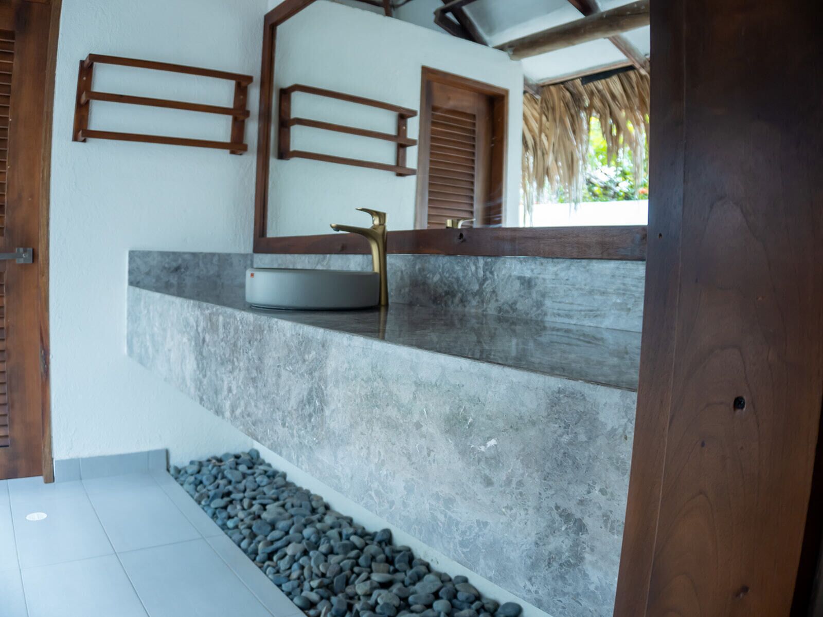 A bathroom with a stone sink, wooden shelves, and a thatched roof visible through the window.